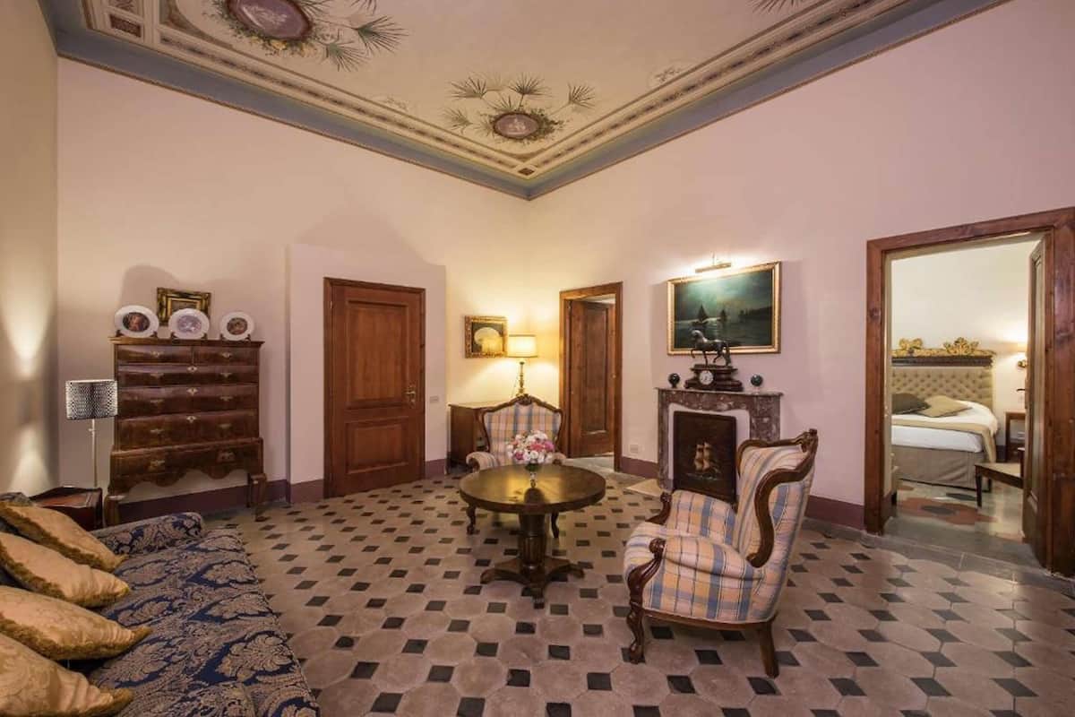 Una habitación con piso a cuadros, una mesa redonda con sillas, un aparador de madera con cuadros enmarcados y una cama al fondo.