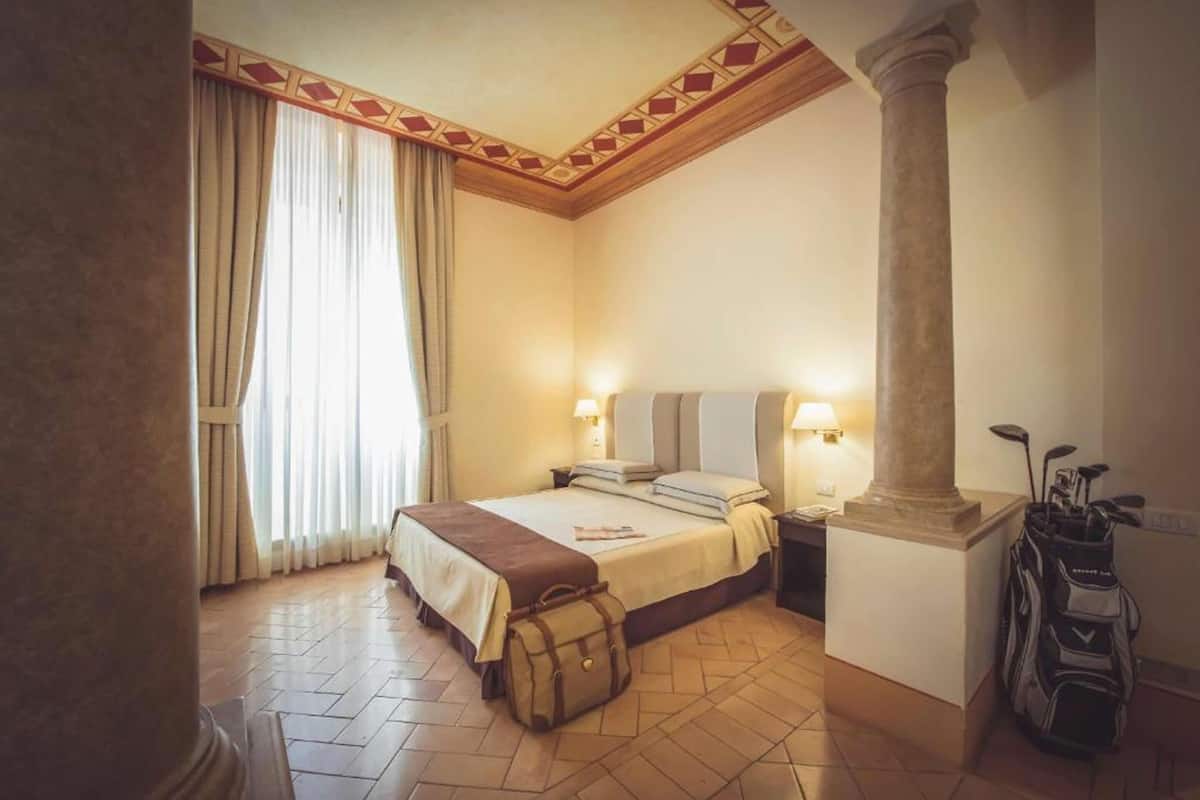 Una habitación de hotel con cama, mesitas de noche, lámparas, un pilar grande y un bolso de golf.