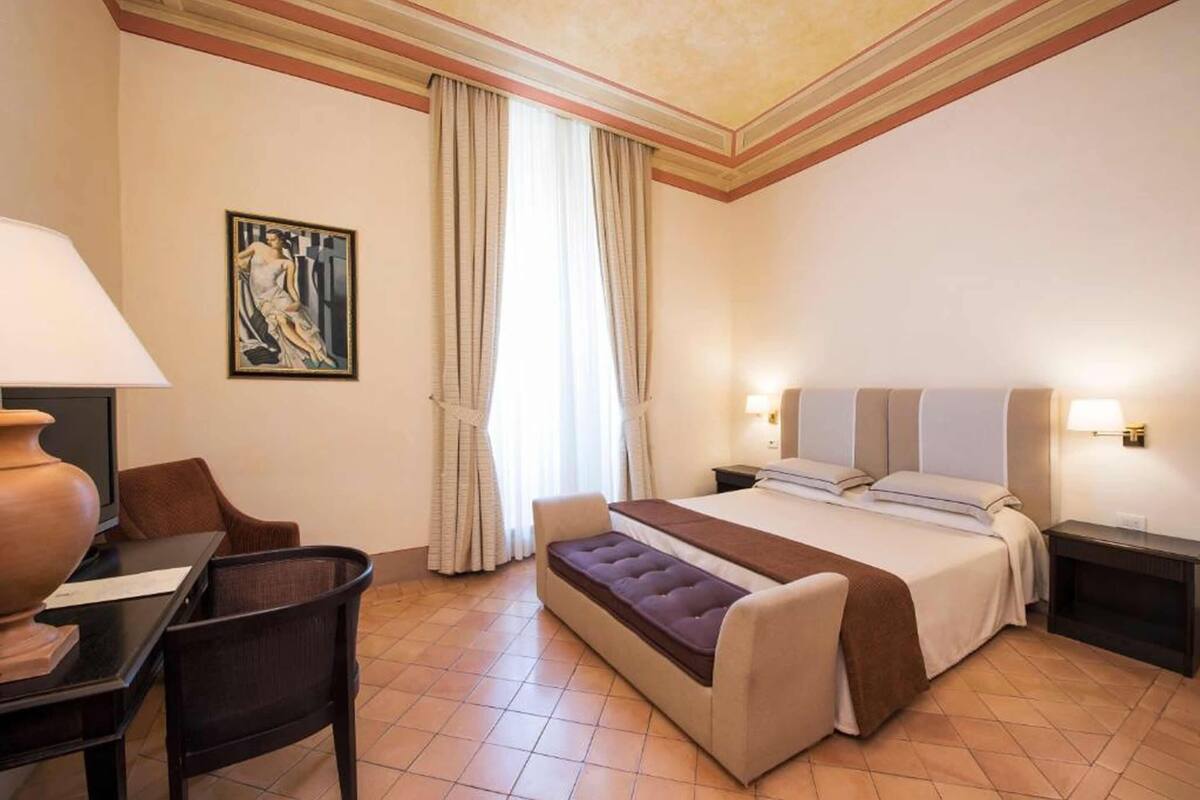 Habitación de hotel con cama, mesitas de noche, un escritorio con silla, un banco, un ventanal con cortinas y un cuadro en la pared.