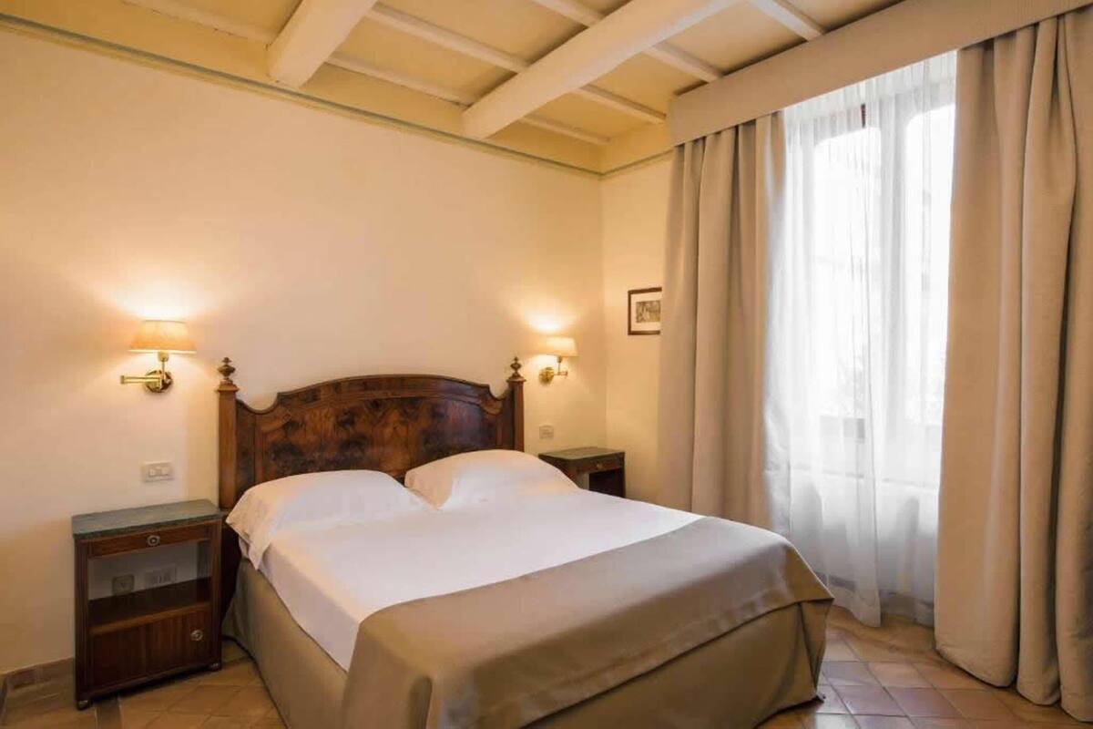 Un dormitorio con cabecera de madera, dos mesitas de noche con lámparas, una cama con ropa blanca y un ventanal con cortinas.