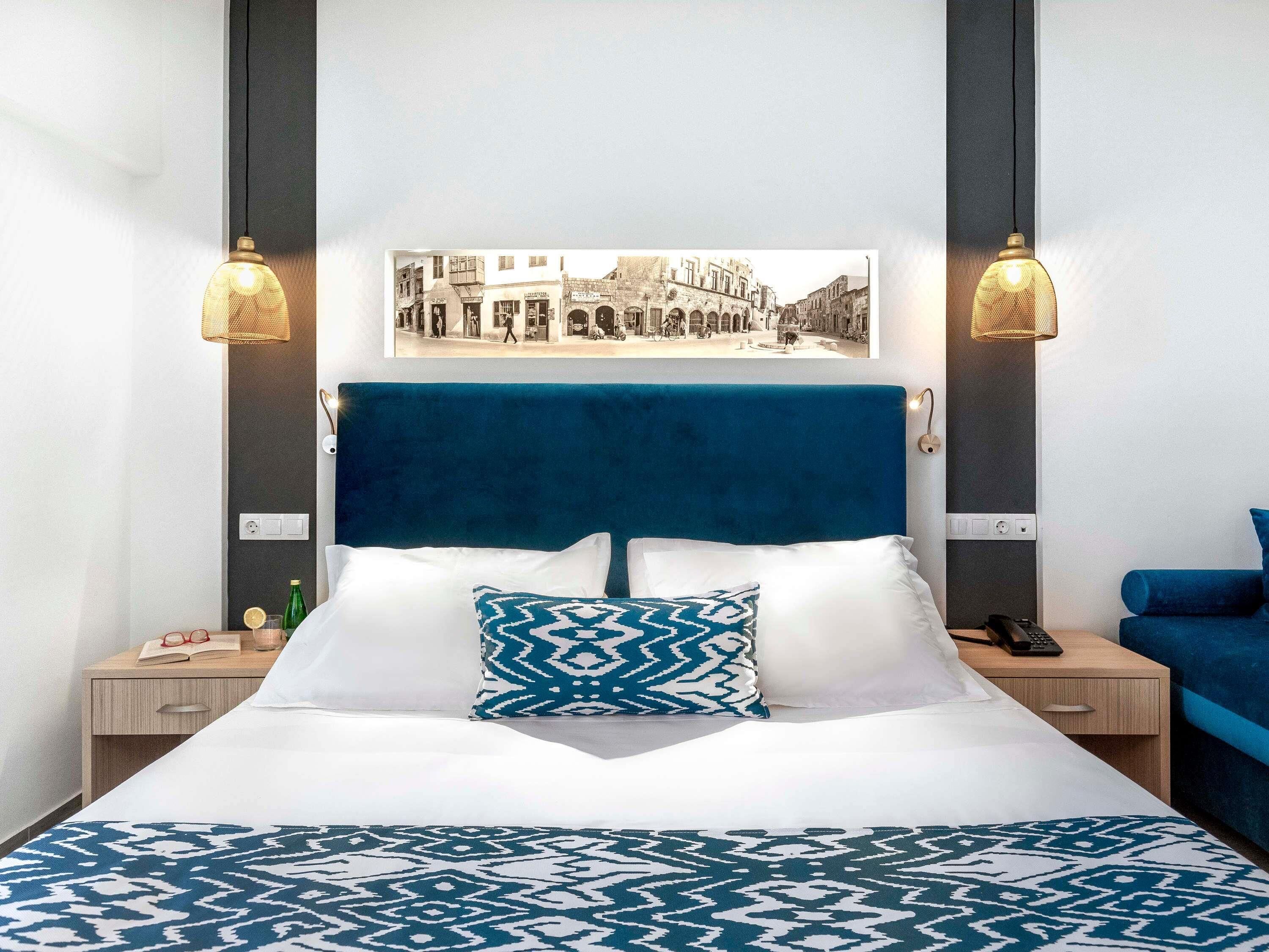 A hotel room with a large bed, a blue headboard, a cityscape picture on the wall, and two bedside lamps.