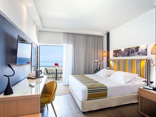 Habitación de hotel con cama, escritorio, silla, televisor, ventana con vistas y un cuadro en la pared.