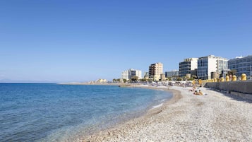 A pebble beach with clear blue water, buildings, and a clear sky.