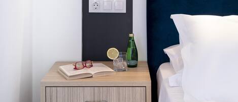 Una cama bien hecha con sábanas blancas, una mesita de noche con un libro, gafas, un vaso y una botella.