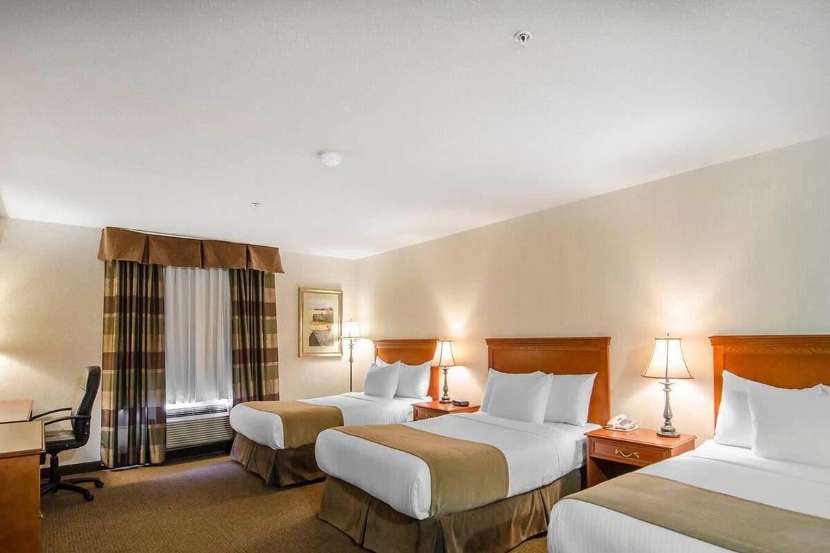 A hotel room with three beds, a desk, a chair, a window with curtains, and a wall-mounted picture.