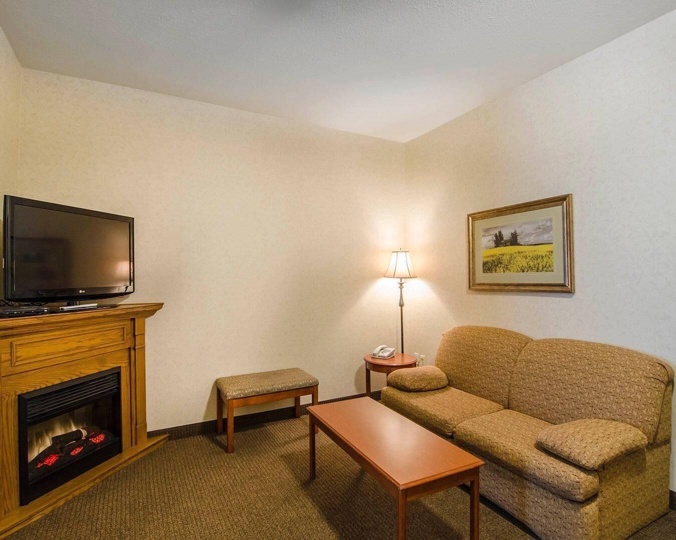Quarto de hotel com televisão, lareira, sofá, mesa, luminária e um quadro na parede.