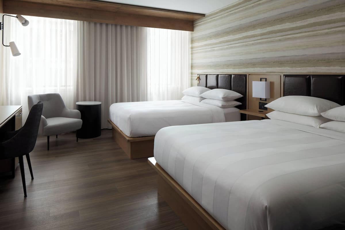 A hotel room with two beds, a desk, a chair, a lamp, and a view of the outdoors through large windows.
