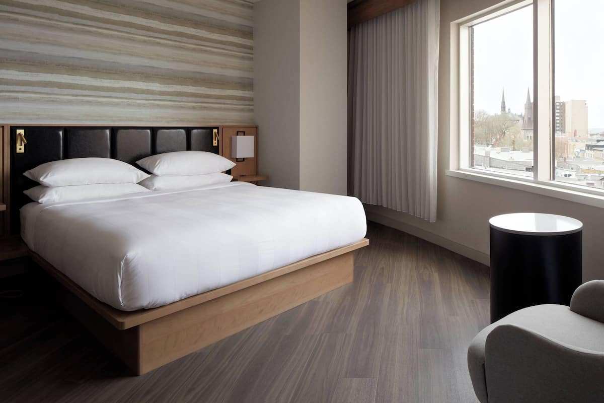 A modern hotel room with a bed, a nightstand, a chair, a window with a city view, and a large window blind.