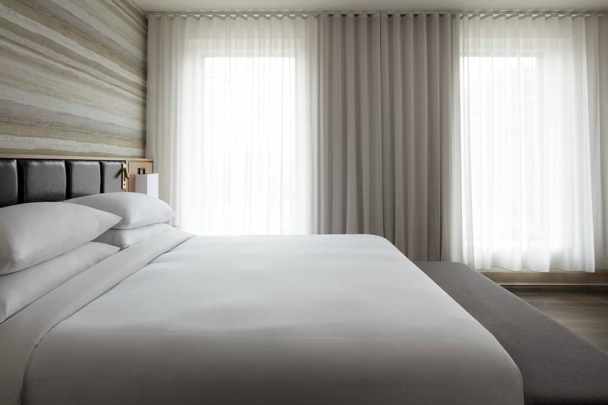 A modern hotel room with a large bed, a headboard with a light fixture, and sheer curtains.