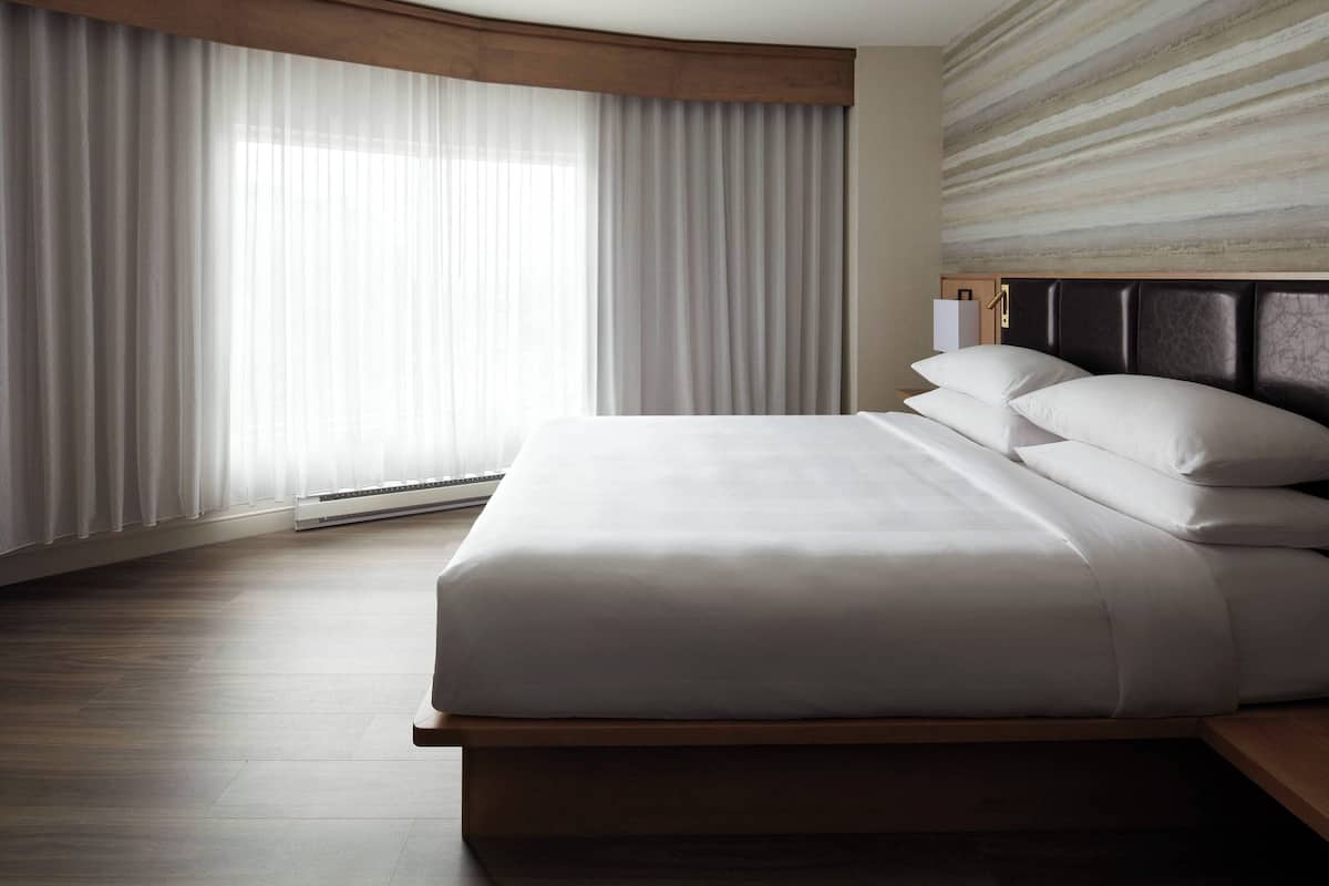 A modern hotel room with a large bed, wooden headboard, and sheer curtains.