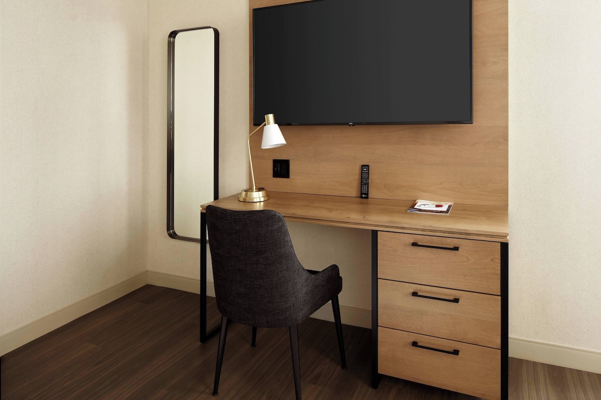 A hotel room with a wooden desk, a black chair, a white desk lamp, a black remote, and a blackboard on the wall.