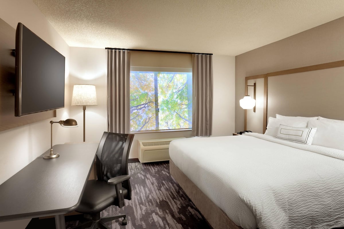Quarto de hotel com cama, escrivaninha, cadeira, televisão, luminária e janela com cortinas.