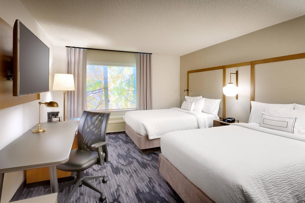 Quarto de hotel com duas camas, uma mesa, uma cadeira, uma televisão e uma janela com cortinas.
