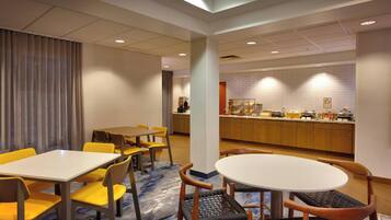A hotel dining area with tables and chairs, a kitchen in the background, and a patterned carpet.