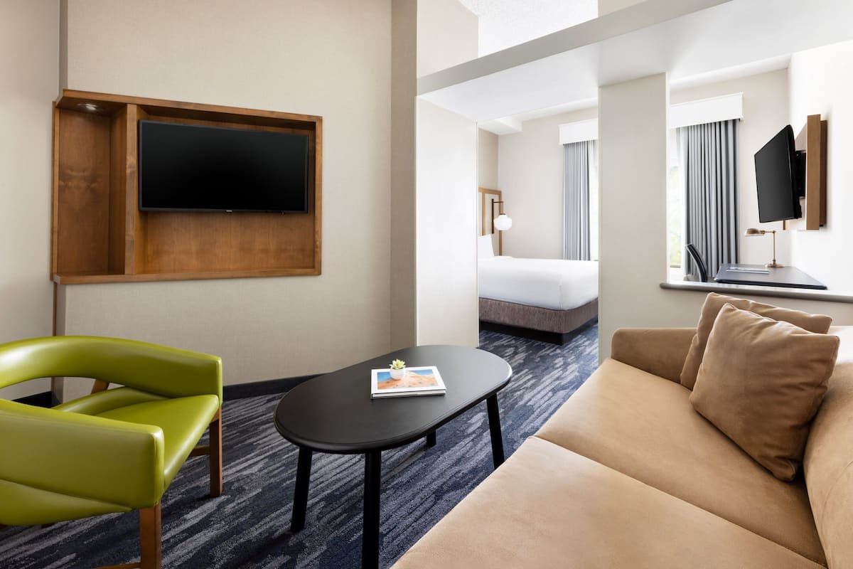 Quarto de hotel com televisão de tela plana na parede, uma pequena mesa preta com uma revista, uma cadeira verde, um sofá bege e uma cama com janela e cortinas.