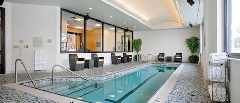 A large indoor swimming pool with clear water, tiled flooring, and a seating area with chairs and a plant.