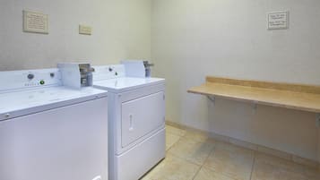 A laundry room with two front-loading washers and dryers, a shelf with a towel rack, and two signs on the wall indicating the location of the front desk and emergency contact information.