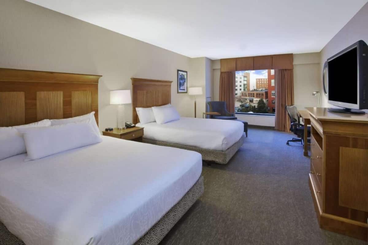 A hotel room with two beds, a desk with a computer, a TV, and a window with a view of buildings.