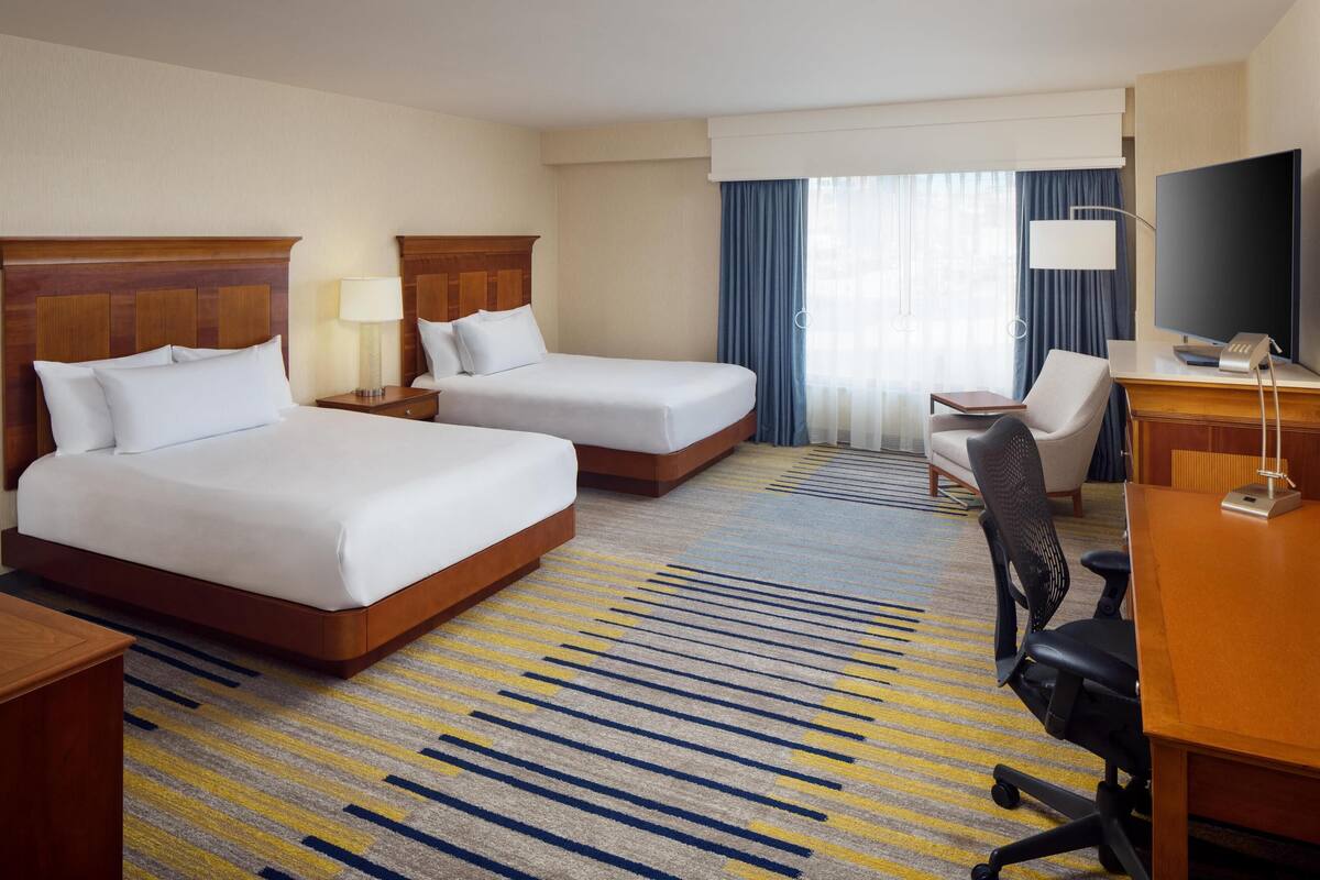 A hotel room with two beds, a desk, a chair, a TV, and a large window with curtains.