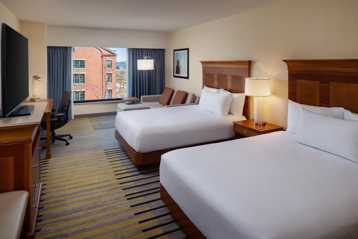 A hotel room with two beds, a desk with a computer, a chair, a lamp, and a view of a building through large windows.