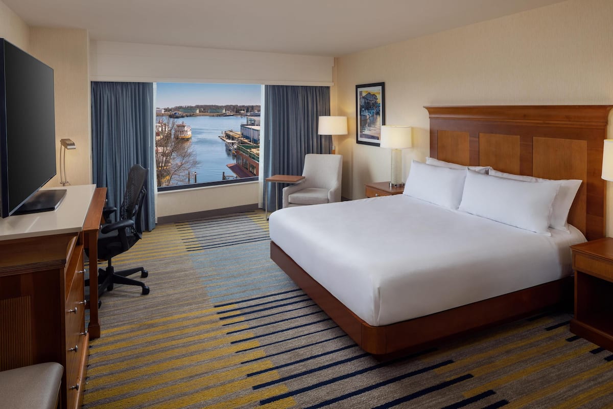 A hotel room with a large bed, a desk, a chair, a TV, and a view of a body of water.