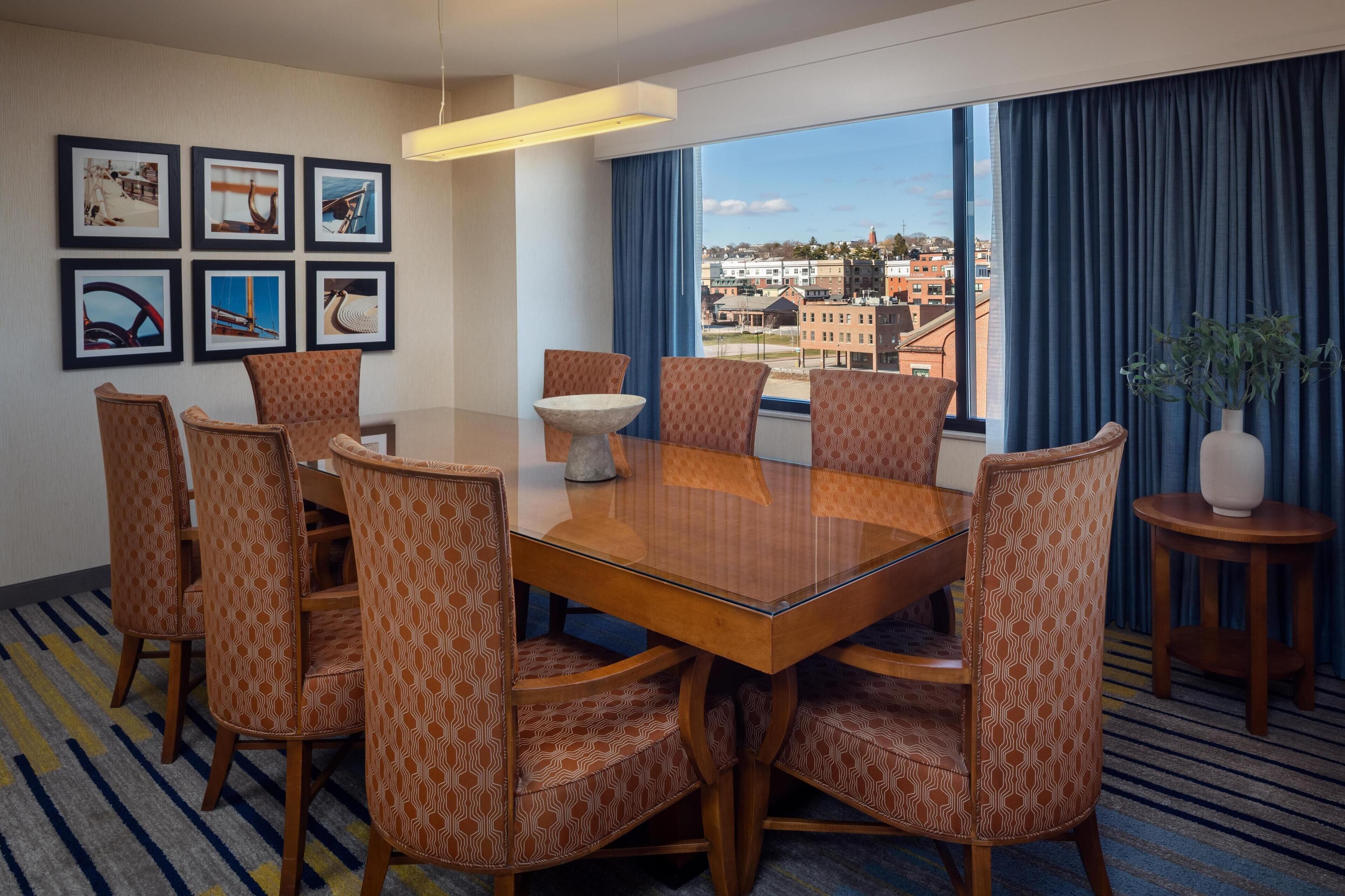 A hotel room with a large window, a dining table with chairs, and framed pictures on the wall.