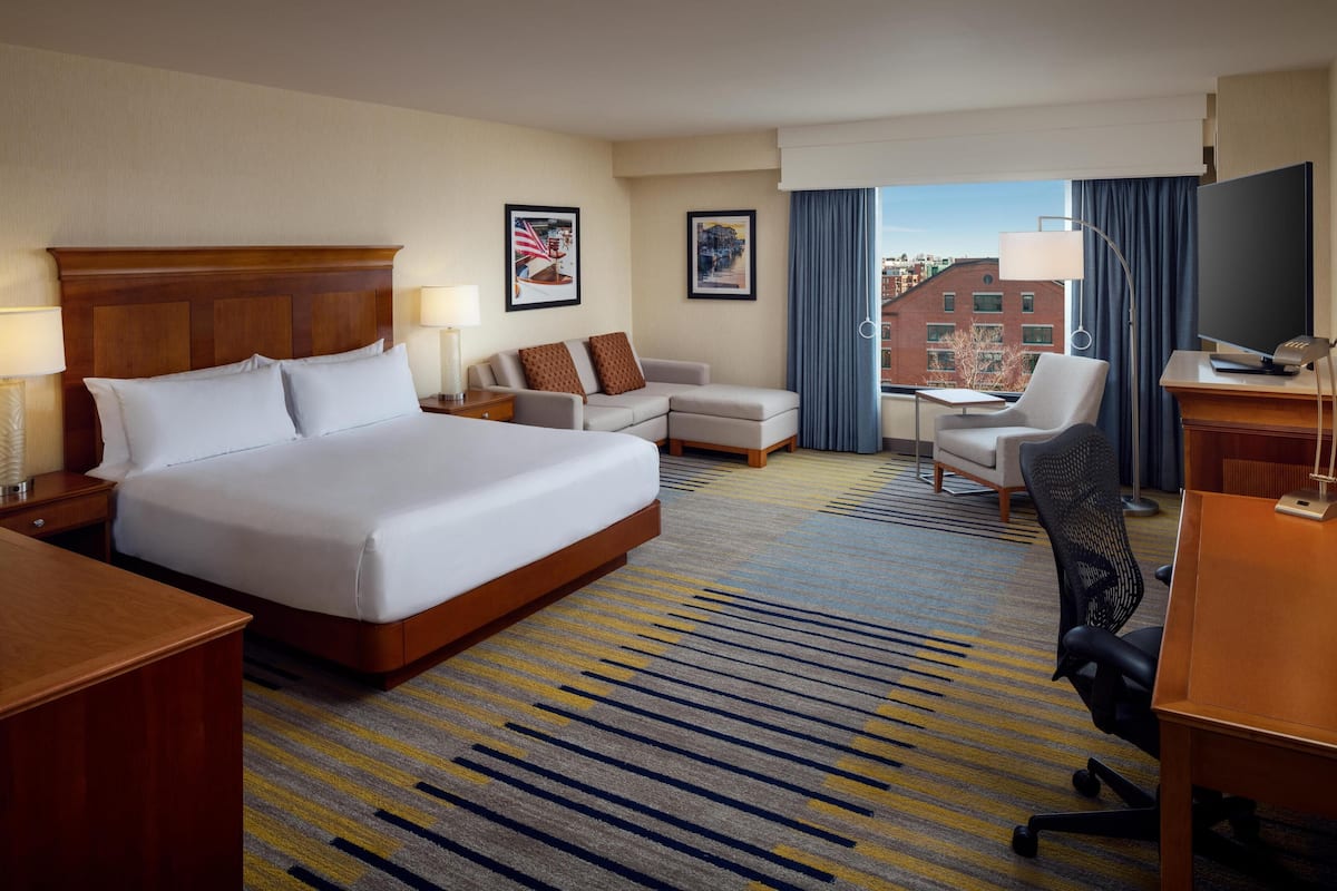 A hotel room with a large bed, a sofa, a desk, and a chair. There are two framed pictures on the wall, a window with curtains, and a TV on a stand.
