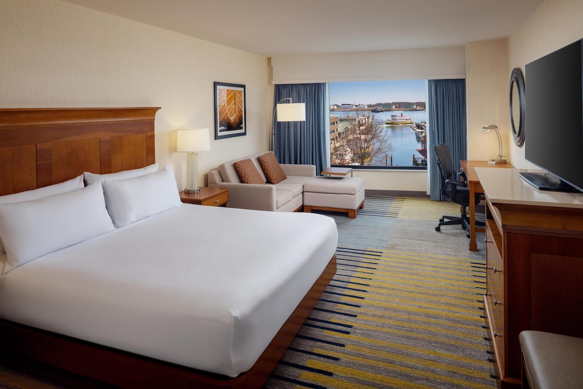 A hotel room with a large bed, a sofa, a desk with a TV, and a view of a body of water and buildings.