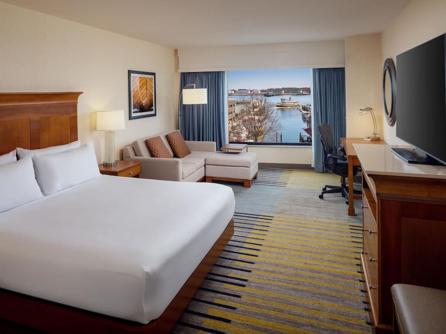 A hotel room with a large bed, a sofa, a desk with a TV, and a view of a body of water and buildings.