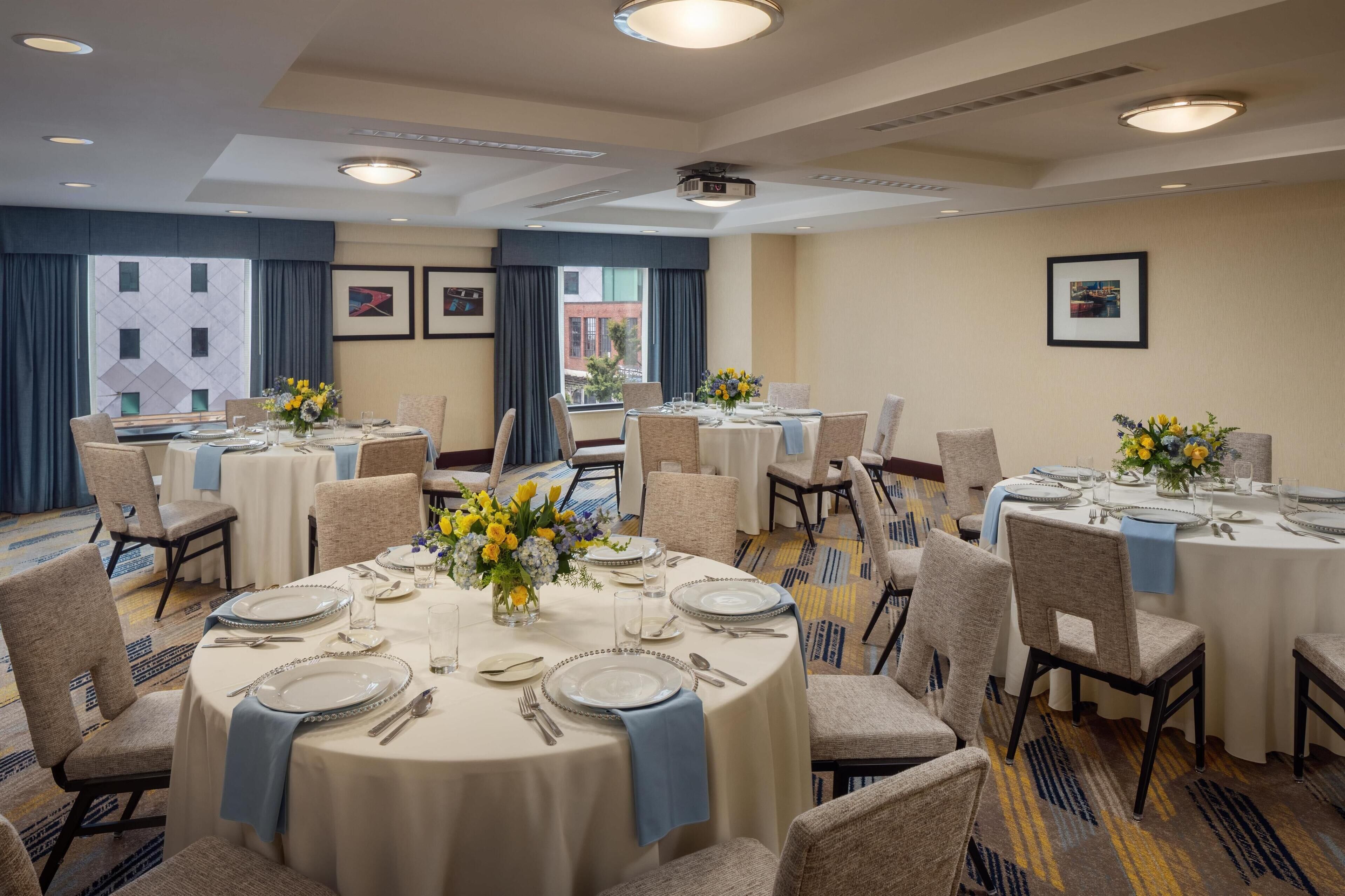 A hotel dining area with round tables set for four, each with white tablecloths, blue napkins, and floral centerpieces.