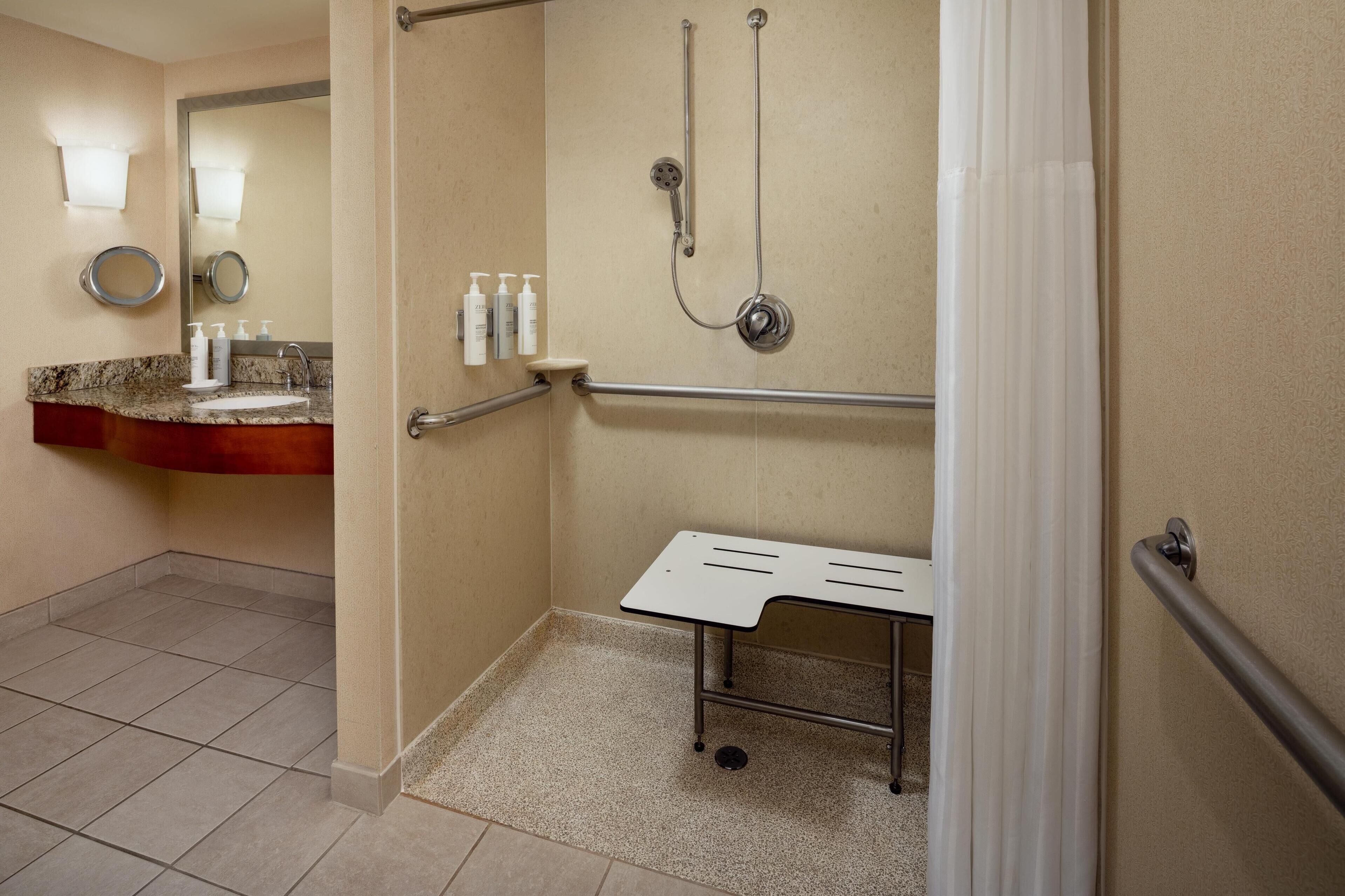 Une salle de bain équipée d’une douche, de barres d’appui, d’un lavabo avec distributeurs de savon et d’un miroir.
