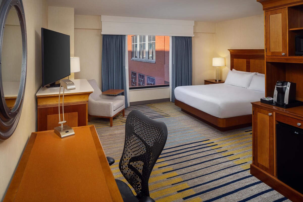 A hotel room with a bed, desk, chair, television, and a view of a building through the window.