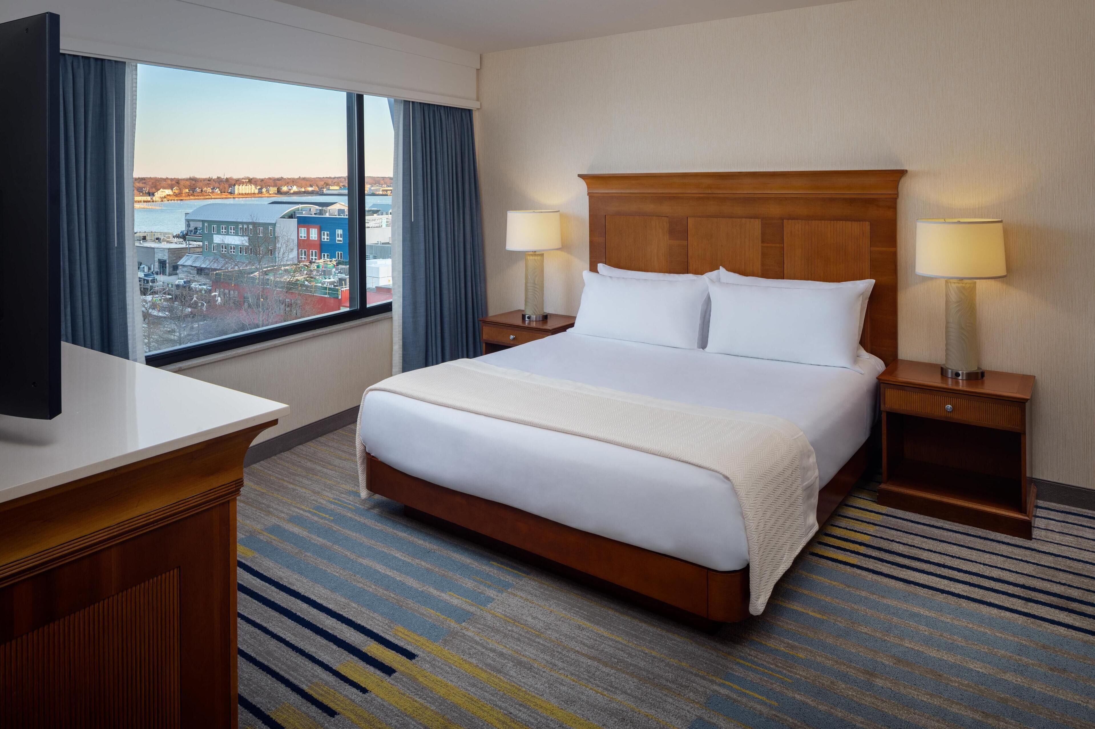 Une chambre d’hôtel avec un grand lit, deux tables de chevet avec des lampes, une vue sur le paysage urbain par une grande fenêtre et un tapis à rayures.