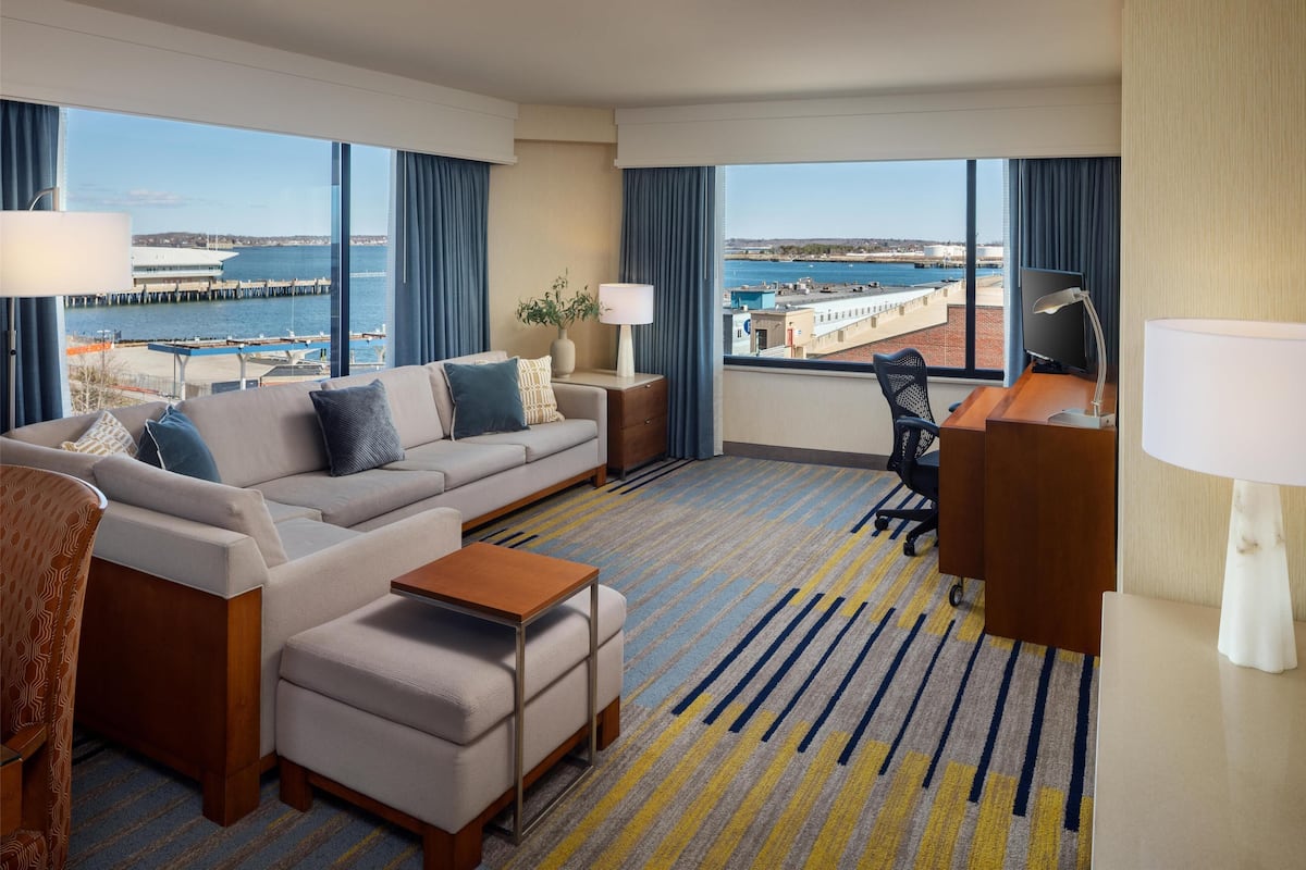 A hotel room with a large window offering a view of a marina and waterfront. There is a sofa, a desk with a chair, and a small table.