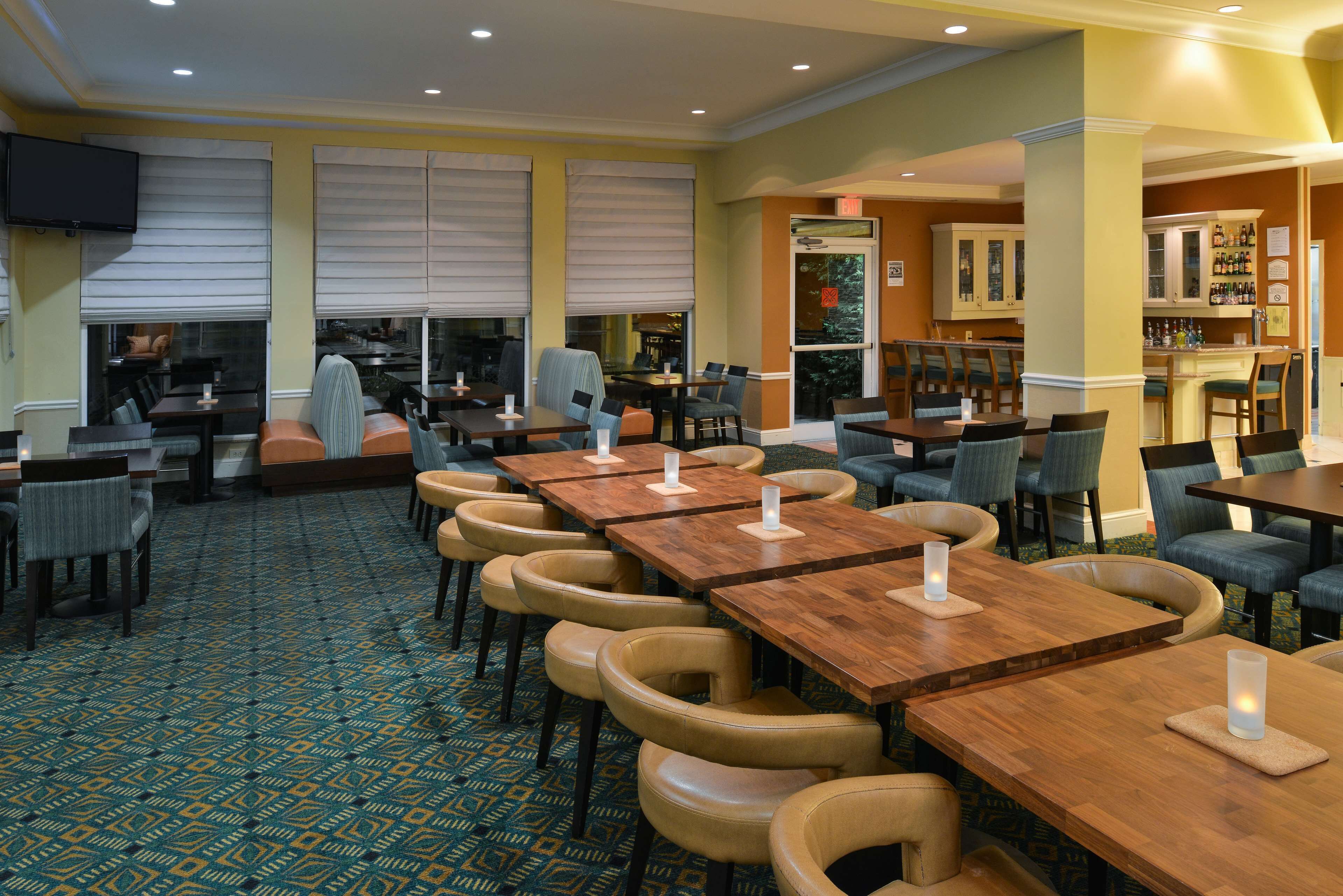 A hotel dining area with multiple tables and chairs, a patterned carpet, and a bar area in the background.