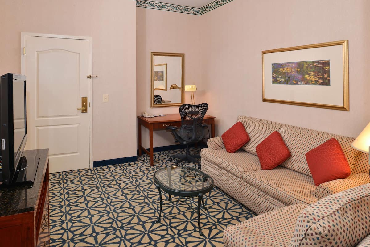 A living room with a patterned floor, a sofa with red cushions, a desk with a chair, a TV, and a framed picture on the wall.