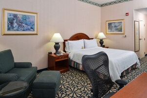 Habitación de hotel con una cama, dos mesitas de noche con lámparas, un sofá verde, una silla negra y alfombra estampada.