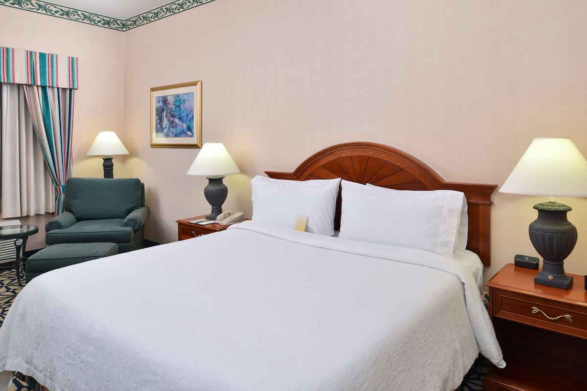 A hotel room with a large bed, two bedside tables with lamps, a chair, a framed picture on the wall, and a striped curtain.