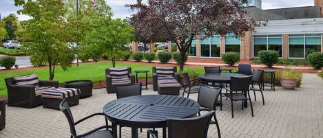 An outdoor patio area with round tables and chairs, surrounded by greenery and a brick pathway.