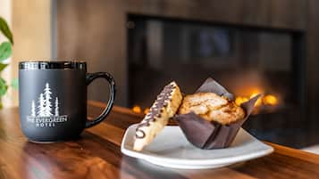 Une tasse noire ornée d’un motif de forêt, sur laquelle on peut lire « The Evergreen Hotel », est posée sur une table en bois. À côté, on trouve une assiette avec deux pâtisseries, dont l’une est nappée de chocolat.
