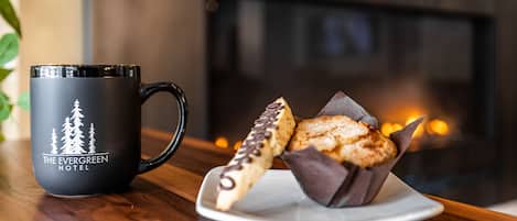 A black mug with a forest design and the text "The Evergreen Hotel" on a wooden table. Next to it is a plate with two pastries, one with chocolate drizzle.