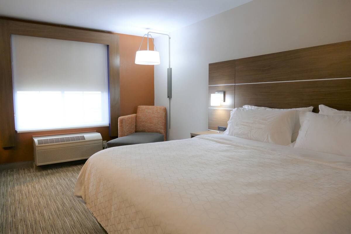 A hotel room with a large bed, a chair, a bedside table, a lamp, and a window with blinds.