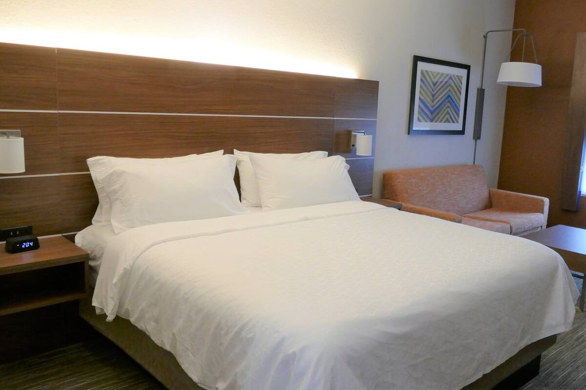 A hotel room with a large bed, a nightstand with a clock, a chair, a small table, a wall-mounted lamp, and a framed picture.