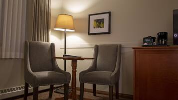 A hotel room with two upholstered chairs, a small wooden table, a floor lamp, a framed picture on the wall, and a coffee maker on a wooden cabinet.