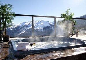 A hot tub with towels and glasses, surrounded by potted plants, overlooking a snowy mountain range.