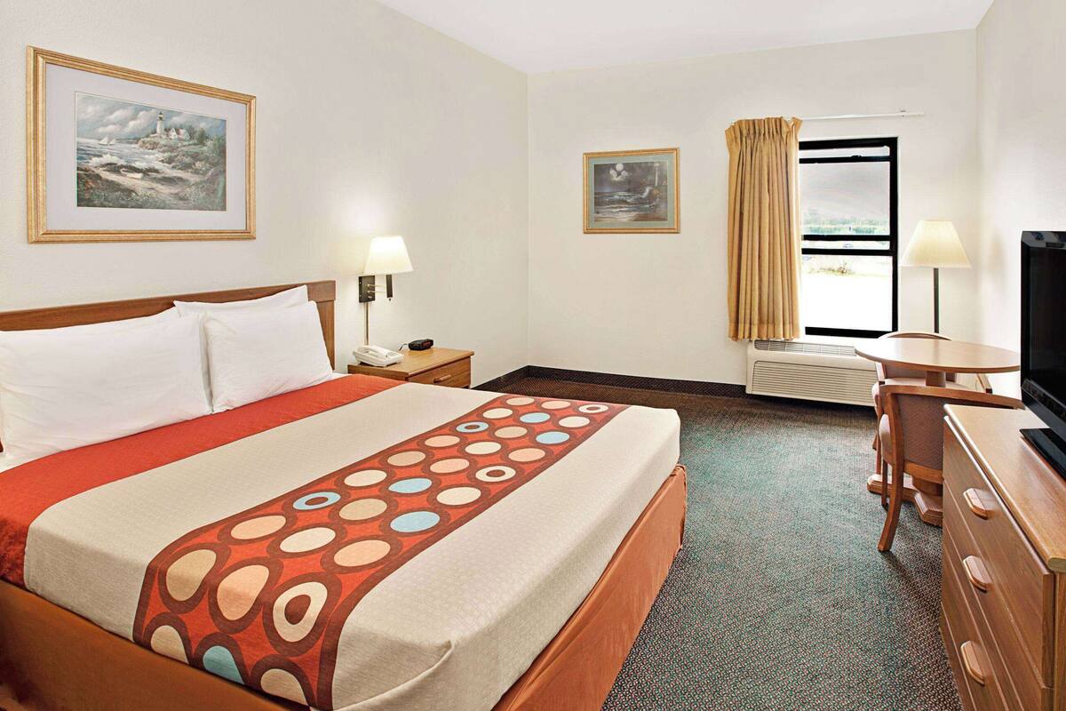 Habitación de hotel con cama, escritorio, televisor, ventana con cortinas y dos cuadros en la pared.