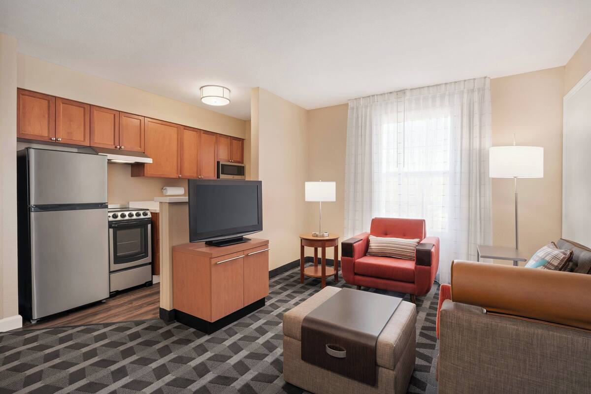 A compact hotel room with a kitchenette, a flat-screen TV, a red armchair, a brown sofa, and a small table.
