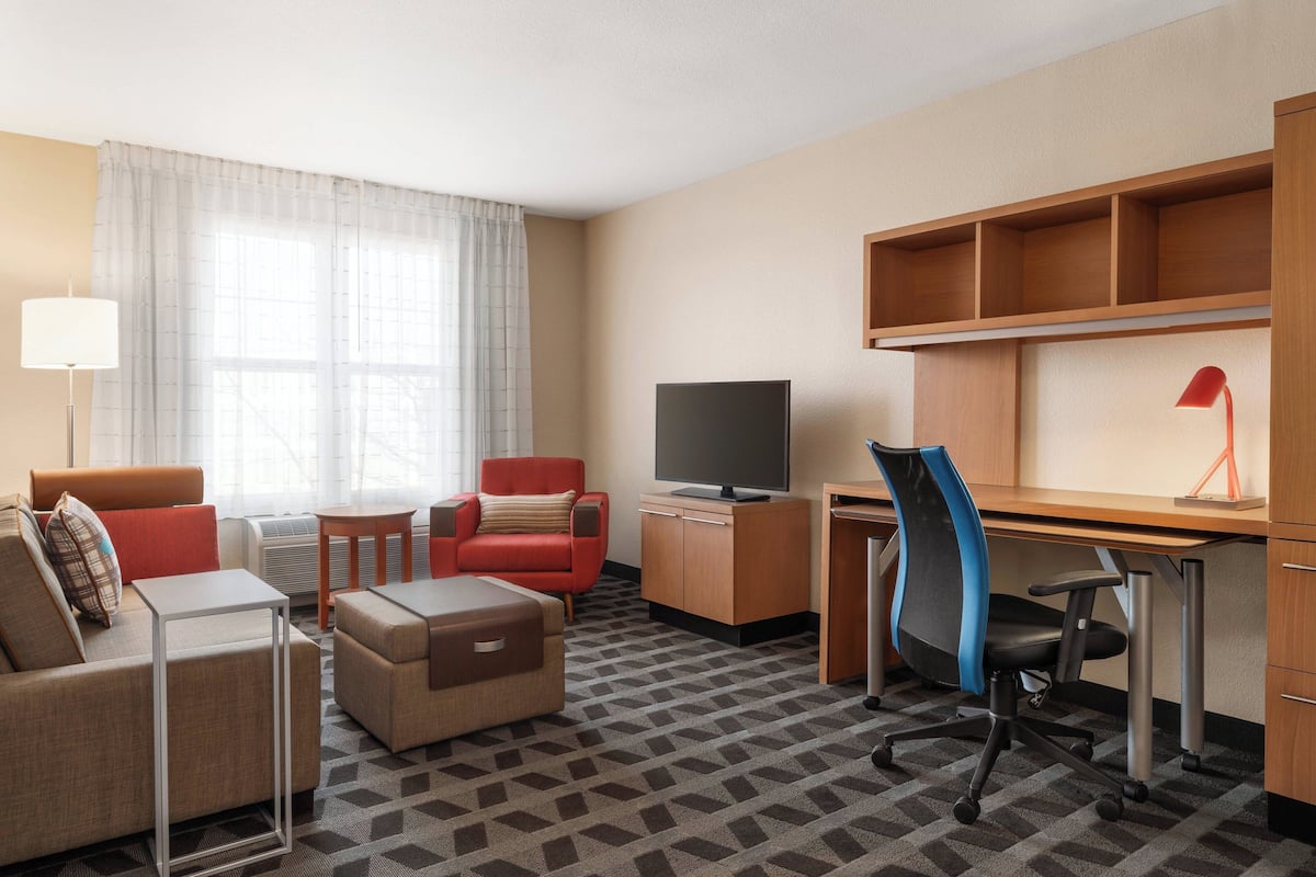 A hotel room with a sofa, armchair, desk, chair, television, and a lamp.