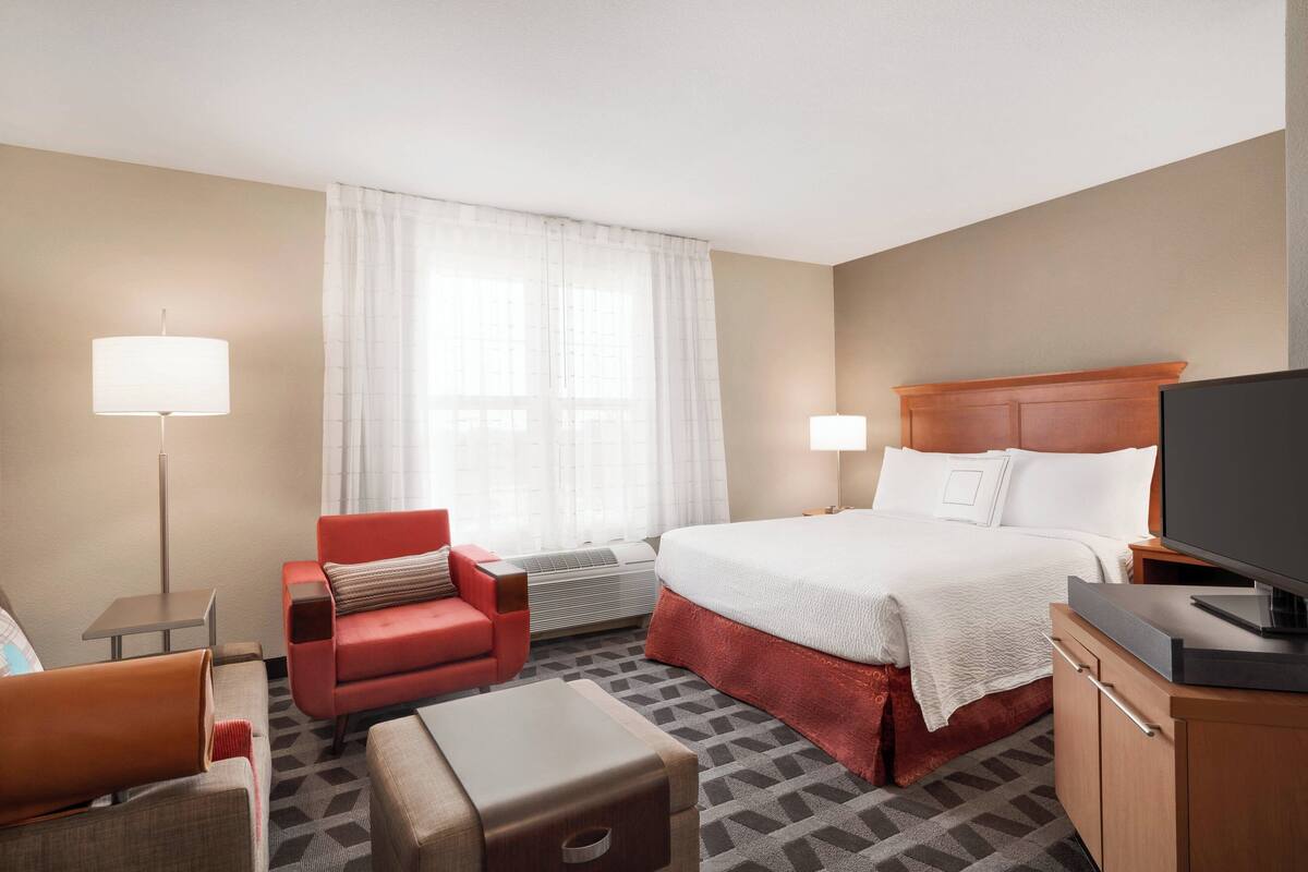 A hotel room with a bed, a red armchair, a sofa, a desk, and a television.