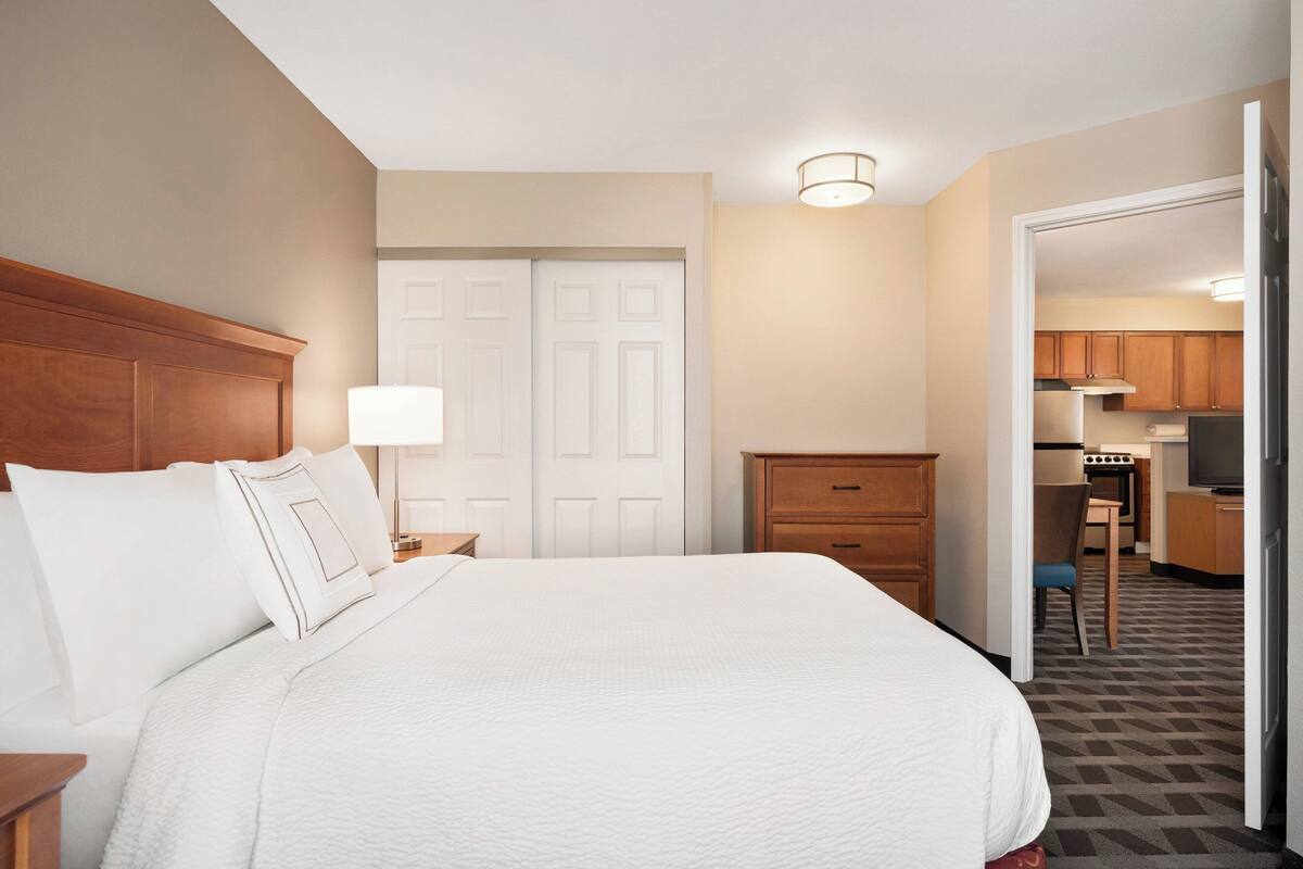 A hotel room with a large bed, a bedside table, a lamp, a wardrobe, and a view into the kitchen area.
